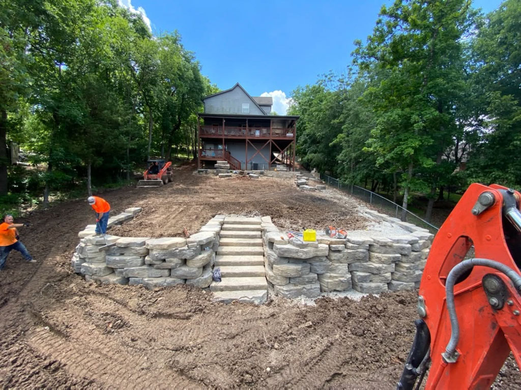 Workers build a stone retaining wall with central stairs in a yard behind a house surrounded by trees.