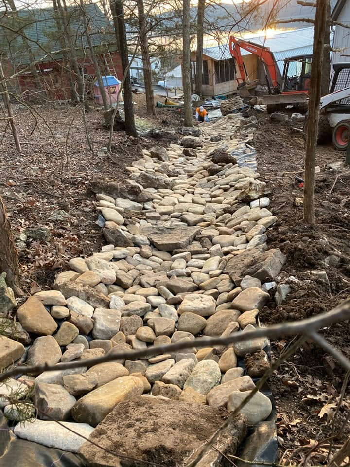 A dry creek bed lined with rocks runs downhill through a wooded area with construction equipment in the background.