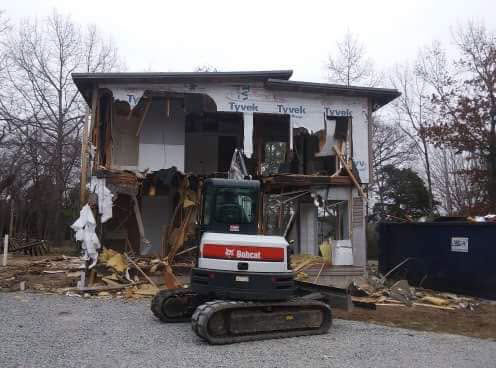 Partially demolished two-story house with exposed walls and a Bobcat excavator parked in front.