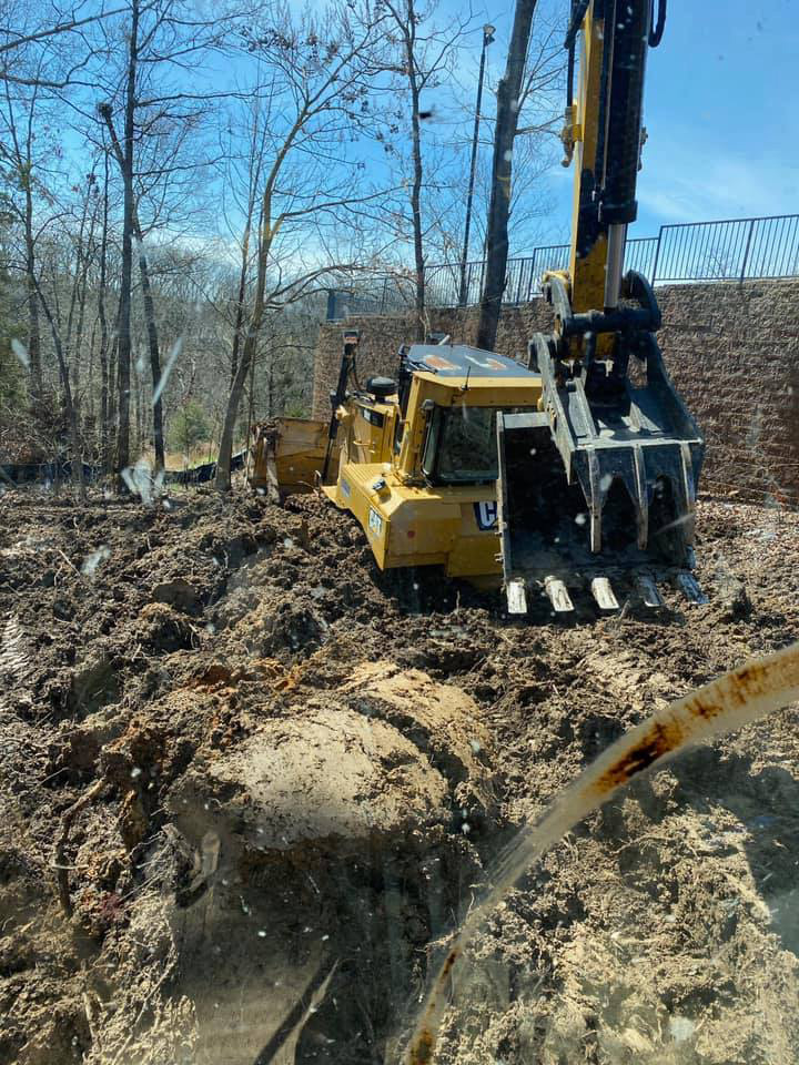 A yellow excavator is stuck in deep mud near trees and a stone retaining wall on a sunny day.